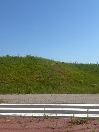 The image captures a clear blue sky above a grassy slope in a rural area. In the foreground, a pedestrian crossing with white stripes is visible on a paved surface. The crossing is bordered by red brick paving with sparse weeds growing between the bricks. Behind the crossing, a gray paved walkway leads up to the grassy incline. The grassy slope is uneven and covered in lush green grass with some patches of dry vegetation. The scene appears to be a public space within the Драчкаўскі сельскі Савет, Belarus, likely a road or pathway adjacent to a green area. There are no people or animals present in the image, and no specific actions are depicted. The environment is natural and calm, suggesting a daytime setting on a sunny day.