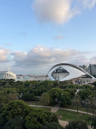 This is an outdoor, eye-level, full shot of the City of Arts and Sciences in Valencia, Spain. The architecture features striking white, futuristic buildings. The primary subject is the Hemisfèric, a building designed to resemble a giant eye, with its distinctive curved structure and the eye-shaped opening. In the foreground, lush green trees and palm trees line pathways and green spaces, indicating a park-like setting. Beyond the Hemisfèric, other structures of the City of Arts and Sciences are visible, including what appears to be the Palau de les Arts Reina Sofía. The sky is partly cloudy with patches of blue, suggesting it is daytime, likely late afternoon or early evening based on the soft lighting. The scene evokes a sense of modern architectural wonder and tranquility within a green urban environment. No people are prominently visible.