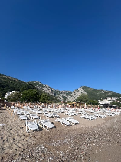 The image shows a sunny day at a beach in Sutomore, Montenegro, with rows of white lounge chairs set up on the sand. In the background, lush green mountains rise behind a coastal town with white buildings. A clear blue sky dominates the upper half of the frame. Several beach umbrellas and a small structure, possibly a beach bar, are visible behind the lounge chairs. A few people can be seen in the distance, enjoying the beach. The sand is a mix of fine particles and small pebbles.