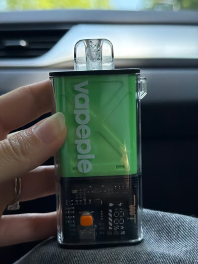 A close-up shot shows a person's hand holding a clear and green electronic device with a digital display. The device, which appears to be a vape, has "vapepie" printed in white on its green body. The digital display on the device shows "100%" with battery and other icons. The background is blurred, with hints of natural light and what appears to be fabric. The person is wearing a ring on their finger. The scene suggests an indoor setting, possibly a car or a room, given the lighting and background elements. The time of day cannot be determined.