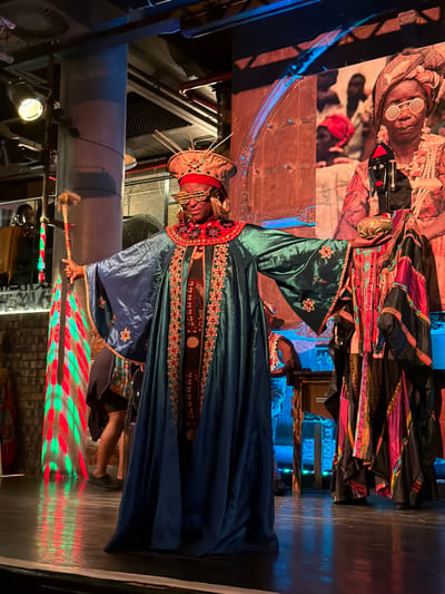 This image captures a person in an elaborate, culturally inspired costume within a dimly lit, theatrical indoor setting, likely in Cape Town, South Africa. The central figure wears a rich, deep blue-green robe adorned with intricate red and gold embroidery along the collar and front opening. A matching broad-brimmed hat or headpiece with similar embellishments covers their head, and they wear distinctive round, white-rimmed sunglasses.

The individual holds a decorated staff or scepter upright in their right hand. The staff's lower portion reflects dynamic red and green diamond-patterned lights onto the dark floor, suggesting an active light display, possibly from a projector or stage lighting.

In the background, a large vertical image, possibly a banner or screen, displays another person in traditional attire and round sunglasses, mirroring the style of the main subject. This backdrop, along with ornate wooden structures and soft blue ambient lighting, creates a vibrant and culturally rich atmosphere. Overhead, industrial pipes and vents are visible, blending with the decorative elements.

The overall scene suggests a cultural performance, an exhibition, or a themed event celebrating African heritage. The artificial lighting indicates an evening or indoor occasion. Faint text "AFRICA" can be seen on a box in the background, reinforcing the cultural context of the setting.