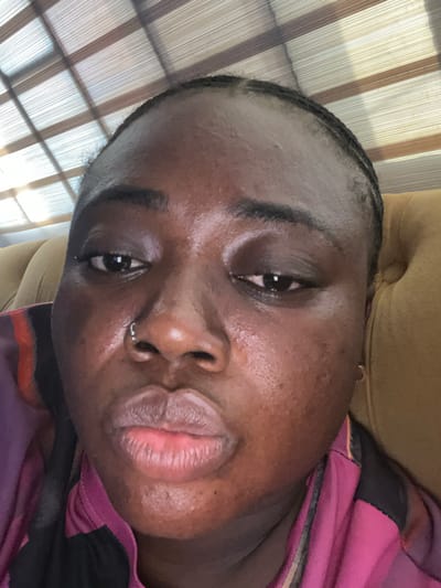 A close-up, rotated portrait of a person with dark skin. The person has dark eyes, dark eyebrows, and a nose piercing. They wear a pink and black garment. The background shows window blinds and a tan-colored surface. Location is unspecified in the image and cannot be determined to be Kubwa, Nigeria.