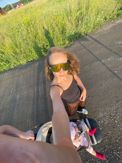 A woman wearing yellow reflective sunglasses, a black tank top, and brown pants is taking a selfie while kneeling next to a stroller on a paved path. A baby in a pink outfit is in the stroller, wearing red socks. The woman's right arm is extended towards the camera, holding a thin stick. A person's arm and shoulder are visible in the foreground. To the left of the path is a grassy field with wildflowers. In the background, houses and trees are visible under a clear sky. The scene appears to be in the village of Podshivalovo, Russia.