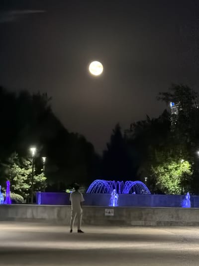 This is a photograph taken at night, likely in Astana, Kazakhstan, under a full moon. The scene depicts an outdoor area with a water fountain illuminated by blue lights. The fountain's streams are captured in motion, creating a dynamic visual. In the background, lush green trees are visible, also lit by ambient light, and the bright full moon dominates the dark sky. A solitary figure, dressed in light-colored clothing, stands near the fountain on a paved walkway. The person's back is to the camera, and their posture suggests a moment of quiet observation. The overall atmosphere is serene and slightly mystical, with the moonlight and artificial lighting creating a unique ambiance. The paved walkway and the modern fountain design suggest an urban park or public space. The time of day is clearly night, indicated by the darkness of the sky and the prominent moon. The weather appears clear, allowing for an unobstructed view of the moon. There is no visible text in the image.