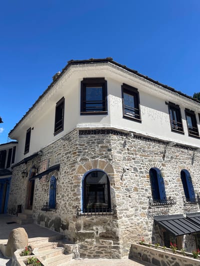 The image shows a close-up, low-angle view of the corner of a stone and white-plastered building in the city of Shiroka Laka, Bulgaria. The building features a tiled roof with decorative white string lights along the eaves. The lower half of the building is constructed from rough-hewn, light-colored stones, while the upper half is smooth white plaster. There are several windows with dark wooden frames. Some of the windows on the stone level are arched and have blue decorative surrounds and wrought-iron grilles. Small flower boxes with red flowers are visible on the ledges below some of these windows. A set of stone steps leads up to a doorway on the left side of the building. The sky above is a clear, bright blue.