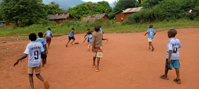 This dynamic image captures a group of young boys playing football on a reddish-brown dirt pitch in Ngalula, Democratic Republic of the Congo. Approximately eight to ten boys, mostly seen from behind, are scattered across the field, their postures indicating active movement in a game.
Many of the boys are wearing football jerseys. One boy in the immediate foreground wears a white jersey with "BENZEMA 9" printed in black on the back. Another boy further back wears a light blue jersey also with "BENZEMA 9." Other blue jerseys are visible, featuring names like "BAKAMBU 17" and "LUKAKU 17." Some boys are barefoot, while others wear sandals or flip-flops. Their expressions and body language convey the typical energy and enthusiasm of children at play.
The scene is set against a backdrop of simple, rural structures. Several single-story houses or huts, constructed with mud bricks or similar materials, have either thatched or corrugated iron roofs. These dwellings are surrounded by dense, lush green vegetation, including numerous trees and bushes, indicating a fertile environment. In the far distance, rolling hills or mountains are visible under a bright, overcast sky. The overall lighting suggests it is daytime, likely late morning or afternoon. The lack of harsh shadows implies a cloudy day. The image vividly portrays a moment of unadulterated childhood joy and activity within a humble, verdant community setting.