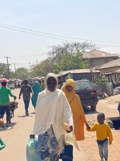 The image presents a busy street scene within Zaria, Nigeria, under bright, clear daylight. In the foreground, an older woman with a medium complexion walks towards the viewer's left. She wears a white hijab, a white patterned long-sleeved top or shawl, and a blue and white patterned skirt. She carries a light blue bag in her left hand and a smaller, light-colored wrapped object in her right. To her immediate right and slightly behind, a younger woman with a similar complexion wears a yellow hijab and a long yellow dress. She holds the right hand of a young, dark-skinned child who wears a bright yellow long-sleeved top and blue jeans, walking on the unpaved dirt shoulder to the far right.
The street is an unpaved, reddish-brown dirt road populated by numerous pedestrians. On the far left, a man in a green shirt is visible pushing a handcart. Other individuals are distributed throughout the mid-ground, including one person in a teal garment. Along the right side of the street, a parked dark-colored hatchback sedan, possibly maroon, is visible behind the foreground figures. Beyond the vehicle, a row of single and two-story buildings line the street, many featuring corrugated metal roofs. A prominent two-story building in the mid-ground exhibits a light green facade with reddish-brown window shutters and a corrugated roof. Sparse trees with dry branches and some green foliage are present in the background. Multiple overhead utility wires are visible against a clear, light blue sky.