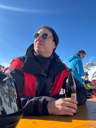 A male individual is seated outdoors at a wooden table under a clear blue sky in Klösterle, Austria. He wears a black beanie, clear-framed circular sunglasses reflecting snow, a black neck gaiter, and a red and black ski jacket. His right hand holds an open brown glass bottle with a "Stiegl" label, resting on the wooden table. His left forearm is also on the table. He is looking upwards and to his left, with a neutral expression. In the midground, another male in a bright blue jacket stands with his back partially to the camera. Further in the background, snow-capped mountains are visible. Other individuals are partially visible at adjacent tables. A black, reflective object, possibly a ski helmet or visor, is on the table in the left foreground. The scene is brightly lit by sunlight.