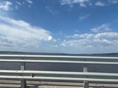 The image shows a view from a bridge overlooking a wide body of water, likely a river or a lake. The water is dark and choppy, reflecting the blue sky and white clouds above. In the distance, the shoreline is visible, lined with trees and a few scattered buildings. The bridge itself has a metal guardrail in the foreground, with multiple horizontal bars. The sky is bright blue with scattered white cumulus clouds, some of which are illuminated by sunlight. The overall scene suggests a sunny day. The prompt specifies the location as Сапуголи, Russia, however, without further information or distinctive landmarks, it is impossible to definitively confirm this specific location from the image alone.