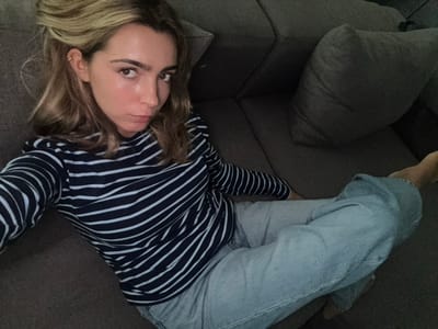 A female individual is depicted in a selfie, seated or reclining on a gray fabric sectional sofa in an indoor setting located in Cidade da Maia, Portugal. She has blondish-brown hair, styled partially swept back, revealing a metallic ear cuff on her left ear. Her facial features include prominent eyebrows and a slight pout. She is wearing a long-sleeved shirt with alternating navy blue and white horizontal stripes and light-wash blue jeans. Her feet are bare, with one visible resting on the sofa. A single gray fabric cushion is present on the sofa. The environment is dimly lit.