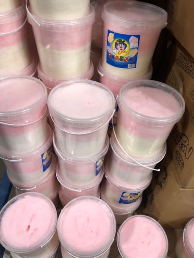The image shows a close-up, high-angle view of several large, clear plastic buckets filled with pink and white cotton candy. The buckets are stacked in multiple rows, with some fully visible and others partially obscured. The cotton candy is fluffy and appears to be layered within the buckets, with the pink layer on top and the white layer beneath. One bucket in the middle row has a colorful label featuring a cartoon clown character giving a thumbs-up, along with images of cotton candy and text in a foreign language, likely Russian. The background consists of cardboard boxes, suggesting a retail or storage environment. The scene is indoors, with artificial lighting. There are no people visible in the image. The setting is identified as Petropavlovsk-Kamchatsky, Russia, implying this is a commercial display or inventory of confectionery products.