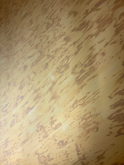 This image presents a close-up view of a textured decorative surface, located in Ihtiman, Bulgaria. The surface exhibits a primary golden-yellow hue, upon which irregular, reddish-brown brushstrokes or patterns are applied. These patterns predominantly trend diagonally from the upper-left to the lower-right, creating a distressed or faux wood grain effect. Scattered areas, particularly towards the middle and lower-left, show subtle lighter, almost white, smudges or streaks within the pattern. The lighting is ambient, highlighting the surface texture without casting sharp shadows. No people, discernible objects beyond the surface, actions, or interactions are depicted.
