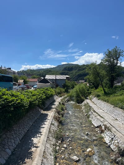 In Dulovine, Montenegro, a clear, shallow stream flows through a channel lined with stone retaining walls on either side. Lush green foliage borders the left side of the channel, with a few parked cars and a blue bus visible behind it. Buildings with tiled roofs are nestled amongst trees on a gentle slope rising in the background. The distant hills are covered in dense green forest, and the sky above is a brilliant blue with scattered white clouds. The overall scene depicts a tranquil, semi-rural setting on a sunny day.