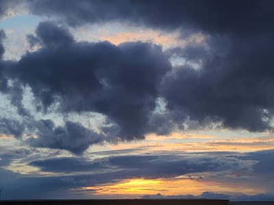 The image presents a dramatic and colorful sky, likely at either sunrise or sunset, over Krasnogorskoye, Russia. The scene is dominated by a diverse array of clouds against a backdrop of varying light.
In the upper and central portions of the frame, large, dark, and dense clouds of grey and deep blue hues are prominent. These clouds appear heavy and textured, with some displaying softened edges while others have more defined, billowing forms. Patches of lighter sky, ranging from pale blue to soft pink and peach, are visible between these darker formations, particularly towards the upper left.
Closer to the horizon, a brilliant and intense glow of golden yellow and orange light indicates the position of the sun, which is either ascending or descending. This radiant light illuminates the edges and undersides of the lower-lying clouds, creating vivid streaks and warm tones that contrast sharply with the cooler, darker clouds above. These lower clouds often appear in elongated, layered formations, some reflecting the intense golden light, while others remain in shadow.
At the very bottom of the frame, a dark, indistinct silhouette suggests a distant horizon, possibly land or a low-lying structure, anchoring the expansive sky. The overall impression is one of dynamic atmospheric conditions and the striking beauty of a transitional moment in the day. No text is visible in the image.