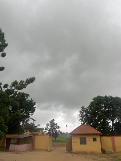 The image depicts a slightly overcast day in Keffi, Nigeria, with a focus on a gated entrance to what appears to be a residential or administrative compound. The sky is filled with heavy, gray clouds, suggesting an impending rain or a generally gloomy atmosphere.
In the foreground, a sandy path leads to the entrance. To the left, a small, open-sided structure with a thatched roof is visible, constructed with wooden posts and pinkish bamboo or reed walls. Adjacent to this is a section of a yellow wall with a red trim, forming part of the compound's perimeter. A tall, slender lamp post with a solar panel stands near this structure.
To the right, a more substantial building with a brown, corrugated metal roof is situated. This building has two windows and a sign with text that is partially legible, reading "NSUK GURUN 21." This building also features yellow walls with red trim, consistent with the other visible boundary walls. A metal gate, partially ajar, provides access through the wall.
The background reveals lush green trees, some quite large and mature, framing the scene. Beyond the compound, a field of green grass stretches into the distance, with more trees dotting the landscape under the muted sky. The overall impression is one of a semi-tropical environment under a threatening sky. There are no people visible in the image.