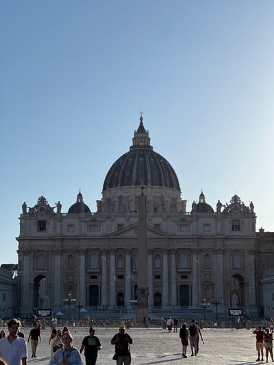 This full vertical shot captures St. Peter's Basilica and a portion of St. Peter's Square in Vatican City, Rome, Italy, under a clear, light blue sky. The grand facade of the basilica dominates the upper background, featuring multiple levels of classical architecture, columns, and a large central dome topped with a cross. Statues line the roofline, and inscribed Latin text, "IN HONOREM PRINCIPIS APOST PAVLV SV BVRGH," is visible above the main entrance area.
In the mid-ground, an ancient Egyptian obelisk stands vertically, partially obscuring the lower facade. The square's paved surface is visible, covered with numerous individuals walking in various directions. Most people are dressed in casual summer attire, and many cast long shadows to their left, indicating the sun is positioned to the right of the frame, likely late in the day. Metal crowd control barriers delineate sections of the square, and a large dark sign with white text is visible near the base of the basilica on the left side.