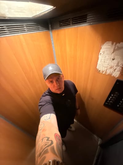 A person wearing a black t-shirt and a grey baseball cap is standing in an elevator with wood-paneled walls. The person is holding a phone to take a selfie, and their left arm, which is heavily tattooed, is visible in the foreground. An elevator control panel with buttons is to the right of the person. The elevator appears to be old and somewhat worn, with a patch of white material on the wall. The overall scene suggests a candid or informal moment captured within the elevator. The location is not discernible from the image itself.