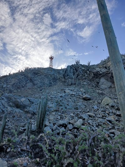 An outdoor, low-angle, medium shot captures a rocky hillside sparsely populated with cacti and other desert vegetation under a cloudy blue sky. The terrain is rough and uneven, with a prominent wooden pole on the right side of the frame and a red and white communication tower situated on the crest of the hill in the background. Several birds are visible in flight against the sky. The scene appears to be in Porlamar, Venezuela, an area known for its arid climate and coastal landscape. The lighting suggests it is daytime, likely late afternoon given the angle of the light.