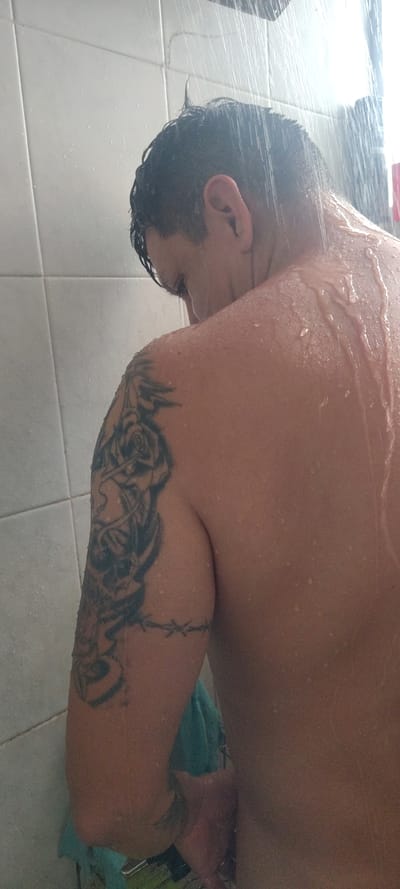 The image captures a person showering from a downward angle, with their back and head visible. The individual's head is tilted forward and down, showing dark, wet hair and one ear. Water streams over their back and shoulders, creating numerous droplets on the skin. A prominent tattoo sleeve is visible on the person's right arm, featuring a black and grey floral design, possibly roses, extending over the bicep, and a barbed wire pattern around the upper arm.

The setting is a shower, indicated by the continuous flow of water and the white tiled floor visible below and behind the person. A light blue towel or piece of fabric is partially visible near the person's lower right side. The overall activity depicted is a person engaged in personal hygiene. No facial expressions are visible, and no specific time of day can be determined from the indoor scene. The provided location context for this scene is Cuernavaca, Mexico. There is no visible text in the image.