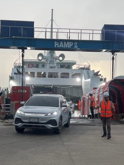 This media file depicts a scene at a ferry terminal, likely in Don Sak, Thailand. The primary subjects are a white car, a large ferry, and several individuals.
The white car is positioned in the foreground, facing towards the viewer's right. Its headlights are illuminated, suggesting it is either preparing to board the ferry or has just disembarked. The license plate is visible and reads "400 8373".
Behind the car is a large white ferry, its side prominently displayed. The ferry appears to be docked, with a ramp structure labeled "RAMP 2" in the lower left corner. Several individuals wearing orange safety vests and hard hats are visible on the ferry and near the ramp, indicating that some sort of loading or unloading operation is in progress. They appear to be workers or crew members.
The setting is a port or ferry terminal with a concrete ground surface. The sky in the background is overcast, suggesting a possibly cloudy day or early morning/late evening. The overall atmosphere seems functional and operational, with a focus on the movement of vehicles and people associated with ferry transport. There are no strong emotional cues, and the scene appears to be a standard day at a busy transport hub.
The visible text includes "RAMP 2" on the blue ramp structure and the license plate number "400 8373" on the car. There is also some text on the ferry, partially obscured, but one recognizable segment appears to be "MO 2".