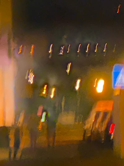 This is a long exposure photograph capturing the blurry streaks of lights from vehicles and possibly streetlights at night. The scene appears to be a street or road in an urban environment, likely Lisbon, Portugal, given the context. The dark background, interspersed with the vibrant, elongated light trails, suggests it is nighttime. The overall mood is dynamic and somewhat abstract due to the motion blur.
The primary subjects are the light trails themselves, which are streaking horizontally and vertically across the frame. These lights are predominantly white, yellow, and red, indicative of headlights, taillights, and possibly neon signs or illuminated buildings in the background. The blurred shapes suggest moving vehicles, possibly cars or trams, though no distinct features of these vehicles are discernible.
There are also some static, more diffuse light sources that appear as blobs, such as a bright white-yellow light in the upper middle of the frame, which could be a streetlight or a building's illuminated window. To the left of this bright light, a blue rectangular shape with white diagonal lines is visible, possibly a sign or part of a building's facade. In the upper right portion, there are suggestions of buildings with orange and brown hues, also blurred by the long exposure. The lower part of the image is a hazy expanse of orange and pinkish light, which might be the illuminated ground or a distant, out-of-focus area.
No people are clearly visible, but their presence is implied by the moving vehicles. The weather cannot be determined, but the slightly grainy texture of the image might suggest some moisture in the air, or it could simply be digital noise. The long exposure technique has rendered the scene as a fluid, almost painterly composition of light and shadow, emphasizing movement and the ephemeral nature of urban night scenes. No discernible text is visible within the image.
