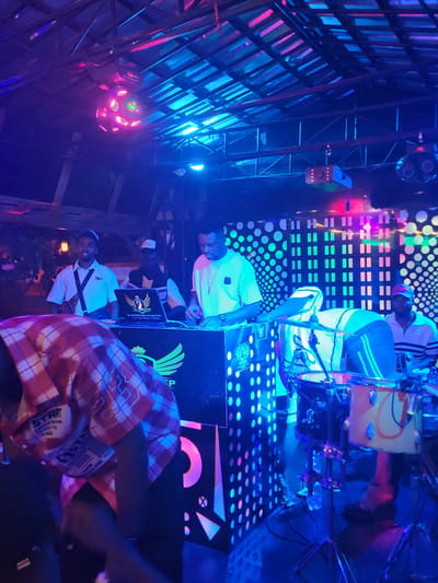Inside what appears to be a nightclub or entertainment venue in Obosi, Nigeria, several individuals are visible under vibrant, colorful lighting. A man in a red plaid shirt stands in the foreground, partially obscuring the view. Further back, two men are positioned at a DJ setup with illuminated panels and equipment. Above them, a man sits atop a structure or platform. The environment is decorated with neon lights and disco balls, indicating a lively atmosphere.