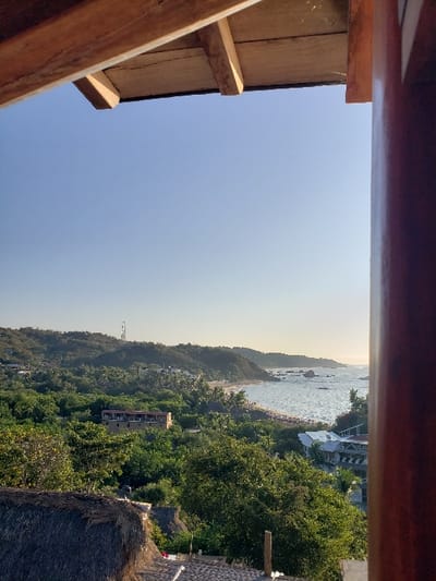 An elevated panoramic view overlooks a coastal section of Mazunte, Oaxaca, Mexico, under a clear, light blue sky. The scene is framed at the top by a wooden ceiling structure with exposed beams and to the right by a partial dark wooden pillar.

In the foreground, the lower left reveals a section of a thatched roof. Adjacent to it, a light-colored ground surface features indistinct objects. Dense green foliage and trees dominate the mid-ground, covering undulating hillsides. Scattered within this vegetation are several buildings, including a prominent multi-story structure with light-colored walls and a red-tiled roof. A tall, slender communication tower is visible on a distant hill to the left.

The background features a curved sandy beach along the Pacific Ocean, populated by numerous small, indistinct human figures and what appear to be beach umbrellas or temporary shelters. The ocean exhibits moderate wave activity, with sunlight reflecting off its surface. Several rocky outcrops are visible both along the shoreline and within the nearshore waters. Hills covered in dense vegetation extend along the coastline, receding into the distance. No specific human actions or interactions are clearly discernible due to the distance.