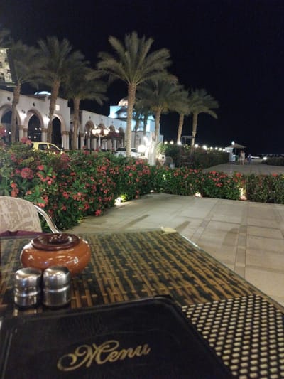The image captures a night scene at an outdoor dining area in what appears to be a resort setting, potentially in an Egyptian location. A table covered with a patterned cloth dominates the foreground, hosting a menu, salt and pepper shakers, and a small ceramic jar. Beyond the table, a paved pathway is lined with flowering bushes and leads towards the beach or waterfront. Palm trees are interspersed along the pathway and in front of a white, arched building, which is lit up. Further back, people can be seen walking near a beachside structure. The environment suggests a tourist-oriented establishment.