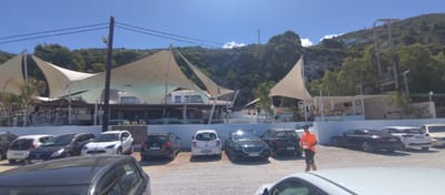 The image captures an outdoor scene under bright, sunny conditions, likely during midday or early afternoon, given the strong light and clear blue sky. In the foreground, a gravel parking lot is filled with numerous parked cars, including a white Peugeot 208 with license plate "AXP 72273", a black Toyota Corolla ("FL11 BXU"), a white Nissan Micra ("EYU 5593"), a black Peugeot 308 ("ZM 124 PE"), a black Mercedes ("CB 9399 KC"), and a black Audi ("G 2721 ZR"). A man wearing an orange t-shirt with "INFO" written on it and dark shorts is walking through the parking lot, holding a white paper or clipboard.

The midground features a modern-looking establishment, possibly a restaurant or beach club, characterized by large, elegant sail-like tensile fabric structures providing shade over an outdoor seating area. The building beneath appears to be multi-leveled with white walls, wooden accents, and white railings. Several people can be seen relaxing or dining in this area. Palm trees and other greenery are interspersed around the building.

In the background, a steep, verdant hillside covered with lush trees dominates the view. Utility poles with power lines are visible running up the hillside, along with what appears to be a large transformer on one set of poles. Scattered white houses with red-tiled roofs can be seen nestled among the trees on the slopes. The scene suggests a popular leisure or hospitality location in a Mediterranean setting, consistent with the Municipal Unit of Sfakiates, Greece.