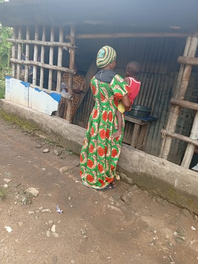 In Owerri, Nigeria, a woman in a colorful green and red patterned dress and a headwrap is holding a child in a red shirt and yellow shorts. They are standing next to a weathered concrete structure. Behind them is a makeshift shelter or building with bamboo framing and corrugated metal walls. Inside, another woman in a patterned dress is visible, seemingly holding a document. A ladder leans against the building. A table with a dark-colored bowl sits inside. The ground is dirt and stone.