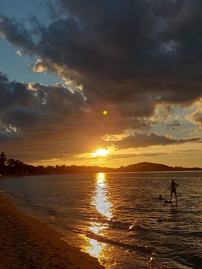 The image depicts a sunset over a beach in Baan Mae Nam, Thailand. The sun is low on the horizon, casting a warm golden glow across the scene. Dark, dramatic clouds dominate the left side of the frame, contrasting with the bright, sunlit sky on the right. The ocean is calm, reflecting the vibrant colors of the sunset, and small waves gently lap at the sandy shore.
In the water, a silhouetted figure is visible, appearing to stand on a paddleboard and holding a paddle. Another smaller silhouette nearby may be another person or an object. The beach itself is sandy, with some vegetation, including palm trees, visible along the shoreline, suggesting a tropical location. The overall mood is serene and tranquil, characteristic of a beautiful sunset. The time of day is clearly sunset, indicated by the low sun and the colors in the sky. The weather appears calm, with the sea being placid.