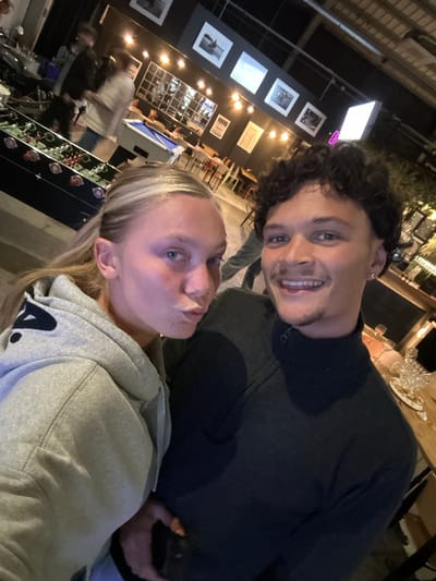 Two individuals, a man and a woman, are captured in a close-up, diagonally oriented photograph within an indoor setting.
The man, positioned at the top right, has dark curly hair, light-colored eyes, and a short beard, smiling broadly at the camera. He is wearing a dark, long-sleeved top with a zipper.
The woman, at the bottom left, has straight blonde hair and blue eyes, looking towards the camera with a slight pout. She is dressed in a light grey hooded sweatshirt with a partial dark logo resembling an "A." visible on the front.
The scene appears to be an indoor commercial establishment, such as a bar, restaurant, or event space, under dim artificial lighting, indicating it is likely evening or night. In the background behind the man, a table is visible with glasses and what looks like a partially eaten dish. The wall behind them is dark, adorned with several framed pictures (some black and white) and a large, grid-like decorative panel. Further in the background, near the woman, a blue pool table or similar game table can be seen, along with blurred figures of other patrons, suggesting a lively social atmosphere. A display rack featuring bottles and some electronic-looking components with knobs is also present near the woman.
The individuals are posing for a photograph, likely a selfie, in a relaxed and social environment in Villeneuve-d'Ascq, France. The man's smile suggests a cheerful mood, while the woman's expression is more neutral.