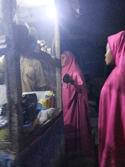 The image depicts a nighttime scene, likely at an outdoor market stall or small shop, characterized by low light conditions and significant graininess. A bright, artificial light source is prominent in the upper center of the frame, casting a strong glare and illuminating a portion of the scene.
There are at least three individuals visible. On the far left, mostly obscured by a vertical structure, a person is seen wearing a light-colored, possibly striped, long-sleeved shirt. Their right arm extends forward towards the center of the frame, suggesting they might be interacting with the stall or another person.
In the center, a person wearing a bright pink hijab and a long, flowing pink garment (likely an abaya or similar dress) is looking intently towards the left, into the stall. Their face is partially obscured by the angle and shadows, but their focus appears to be on items within the stall. This individual is holding a pair of eyeglasses in their left hand, which dangles downwards.
On the far right, partially out of frame, another person is visible, also wearing a bright pink hijab and a matching long garment. This person is facing towards the left, appearing to observe the same activity as the central figure.
The stall itself features a vertical support structure on the left and a visible counter or display area. Above the counter, there's a mesh or wire covering. Inside the stall, behind the person in the light shirt, various indistinct items are present, including a yellow object with purple and white details, possibly a small toy or decorative piece. The overall activity suggests browsing or shopping at the stall.