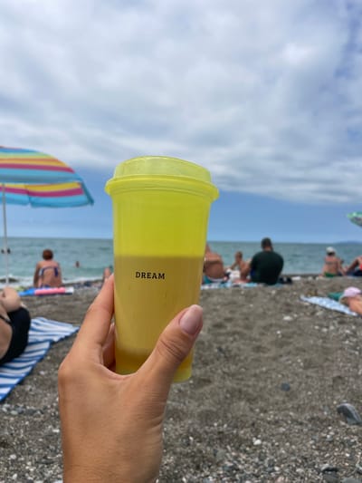 A first-person perspective shot shows a hand holding a yellow plastic cup with a yellow lid. The word "DREAM" is printed in black on the side of the cup. The cup contains a light brown liquid. The background is a beach scene. In the middle ground, people are seated on towels and at the water's edge. An umbrella with colorful stripes is visible to the left, and another umbrella is partially visible to the right. The sea and a cloudy sky form the background. The location is the city of Gagra, Abkhazia.
