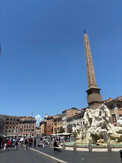 The image depicts a view of Piazza Navona in Rome, Italy, under a clear blue sky. Dominating the right side of the frame is the Fountain of Four Rivers, a Baroque masterpiece by Gian Lorenzo Bernini, with its intricate sculptures and flowing water. Behind the fountain, an Egyptian obelisk, the Obelisco Agonale, rises tall, its ancient hieroglyphs visible against the sky.
The piazza is populated with numerous people walking, standing, and sitting on benches. Some individuals are holding umbrellas, suggesting a sunny day. Buildings with traditional Roman architecture, featuring warm-toned facades and numerous windows, line the perimeter of the piazza. Some buildings appear to be shops or cafes, with awnings visible. The ground is paved with cobblestones. The overall atmosphere is that of a bustling public square on a bright day.