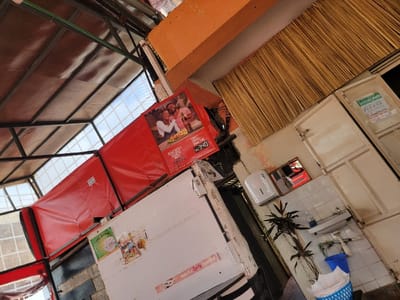 The image displays the interior of what appears to be a local eatery or a covered market stall in Makuyu, Kenya, under natural daylight. The structure features a metal-framed roof with alternating translucent and opaque panels, allowing light to filter in. A prominent red fabric canopy or awning is stretched across the left side of the frame.
On the right side, a functional area is visible, featuring a white tiled wall with a basic washbasin and faucet. Above the sink, a white wall-mounted dispenser, likely for soap or paper towels, is installed, with a small rectangular mirror above it. A potted plant with dark foliage stands on the floor next to the sink, accompanied by a blue bucket and a white and blue woven basket. The upper part of the wall is adorned with a textured, woven material, possibly bamboo or straw, and there are also white panelled cabinets or shutters.
A large red advertisement poster is fixed to the red canopy. It prominently features a smiling man and woman sharing drinks and a plate of food, possibly snacks or fries. The text "KES 340" is clearly visible on this poster, indicating a price in Kenyan Shillings, which aligns with the given location context of Makuyu, Kenya. Other smaller, less legible posters and stickers are also present on a white surface below the red canopy.
No people are physically present in the scene, but the advertisement depicts joyful individuals, suggesting a convivial atmosphere for dining and socializing. The setting has a practical, somewhat rustic aesthetic, typical of local establishments.