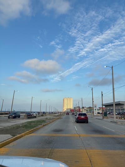 The image shows a road scene in Coatzacoalcos, Mexico. The view is from a car moving along a highway. The road has multiple lanes with yellow dividers. Several cars are driving in the same direction. On the side of the road are street lights and parked cars. Buildings, including a high-rise apartment or office building, are visible along the highway. The sky is blue with scattered clouds.