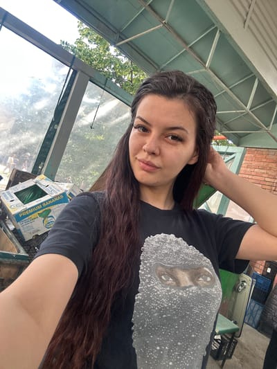 A young woman with long, dark hair poses for a selfie outdoors. She is wearing a black t-shirt with a graphic print of eyes. Her left arm is raised, touching her hair. In the background, there are stacks of banana boxes and various plastic crates, suggesting a market or storage area. A metal and glass structure forms the roof and part of the wall. The scene appears to be in Sliven, Bulgaria, indicated by the presence of what appear to be Bulgarian Cyrillic characters on one of the banana boxes.