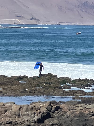 The image depicts a person in a black wetsuit holding a blue boogie board, standing on a rocky shoreline near the ocean. The waves are breaking in the background, and there are several boats visible in the distance. The terrain behind the shoreline appears to be arid and mountainous. Based on the visual cues, the location is likely the coast of Arica, Chile, known for its rocky shores and proximity to desert landscapes. The person appears to be engaged in a water sport, preparing to enter or exit the ocean. The overall scene captures a moment of coastal activity against a backdrop of natural elements.