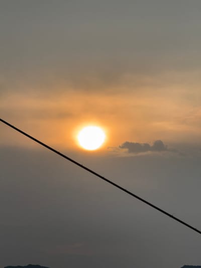 This vertically oriented image captures a hazy sky dominated by a bright, diffused sun. The sun, a luminous white circle with a warm yellow-orange glow emanating from it, is positioned in the lower-central part of the frame, suggesting a low angle in the sky, possibly during sunrise or sunset.
The sky is filled with a soft, warm haze or thin clouds, giving the atmosphere a muted, ethereal quality. A dark, thin, diagonal line, appearing to be a utility cable or wire, stretches across the frame from the bottom-left to the upper-right, crossing in front of the sun. Small, indistinct dark shapes are visible clinging to this line, possibly connectors or insulators.
On the right side of the sun, a cluster of darker, more defined clouds is visible against the lighter sky. The overall color palette is dominated by grays, yellows, and oranges, indicative of atmospheric conditions. No people or specific activities are visible. The image evokes a sense of calm and atmospheric depth. The location context provided is Nam Nha Trang, Vietnam, though no specific geographical features identifying this location are discernable within the image itself. There is no visible text.
