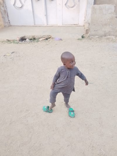 A young child stands in a dirt yard, wearing mismatched green Crocs. The child wears a gray, traditional-style outfit. A white, closed gate is in the background, along with a rough concrete structure. The child appears to be enjoying themselves and looking off to the side.