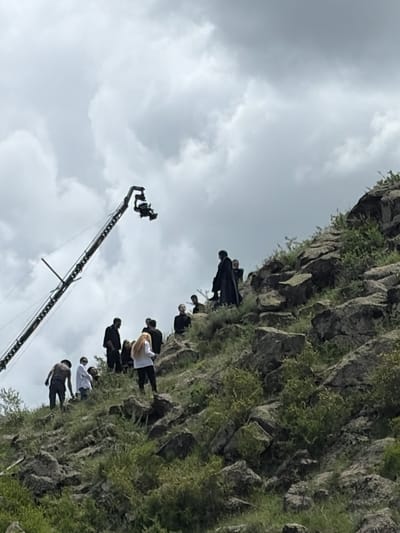 A rocky hillside is shown under a cloudy sky. Several individuals are positioned along the hillside, some wearing dark clothing and at least one person wearing white. A camera crane is present. The scene suggests a film or video shoot. This is taking place in or around Tlik, Armenia.