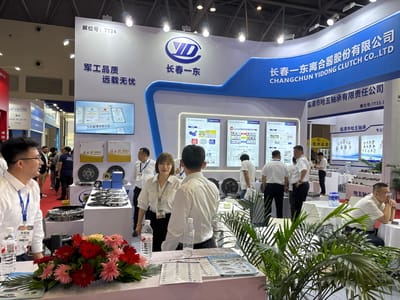 The image depicts an exhibition booth at a trade show. The booth belongs to CHANGCHUN YIDONG CLUTCH CO.,LTD. Several people, mostly dressed in white shirts and dark pants or skirts, are present; some appear to be booth staff engaging with visitors. Products related to clutches are displayed on white tables. Behind the tables are displays with technical diagrams and information about the company and its products. Floral arrangements are visible on the tables in the foreground. Another business called "临清市哈五轴承有限责任公司" or "Linging City Hawu Bearing Co. LTD" is to the right of the Changchun company. A smaller sign reads "展位号:7T33-1," which translates to Booth Number: 7T33-1. The booth's signage states "军工品质远载无忧," translating to "Military Quality, Worry-Free Long Journey," along with "5大管理体系认证," meaning "5 Major Management System Certifications." A smaller sign reads "展位号:7T24" which translated to "Booth Number: 7T24". Another company sign reads 重庆(Chongqing) 2026.05.13-15
7T12 and 四川华车辆有限公司(Sichuan Hua Vehicle Company).