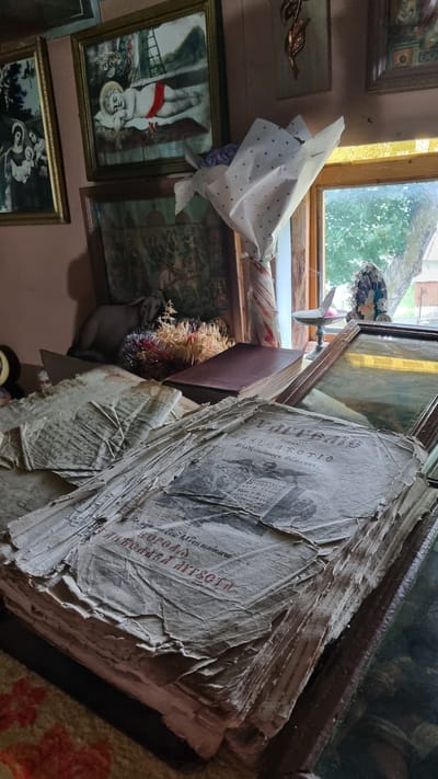 This media file captures a cluttered, indoor scene, likely a room in Vyshka, Ukraine. The focus is on a large, aged stack of what appears to be newspapers or old documents, piled precariously on top of a book. The paper is yellowed and worn, with visible tears and creases, suggesting significant age and frequent handling. Some of the documents have text and images printed on them, though the details are mostly obscured by the stack's condition.
To the left of the stack, a window with wooden frames looks out onto a greenery-filled outdoor environment, suggesting daytime. Sunlight filters through the window, illuminating the room and casting shadows. Several framed artworks hang on the wall. One painting, towards the left, depicts a figure that resembles a child, possibly a religious depiction, with a red sash. Another painting further to the right shows a more complex scene with multiple figures. A bouquet of dried flowers and other decorative elements is tucked beside the stack of papers.
A white wrapping with polka dots covers a portion of something on the windowsill, potentially a gift or another item. The overall impression is one of accumulated possessions, perhaps in a storage area or a room filled with historical items. The lighting suggests it is daytime, and the view from the window hints at a natural, possibly rural, setting outside. No specific activity or emotion is clearly depicted, but the aged nature of the objects could evoke a sense of history or nostalgia. Some text is visible on the topmost newspaper, including what appears to be Cyrillic script, but it is too small and blurry to read clearly.