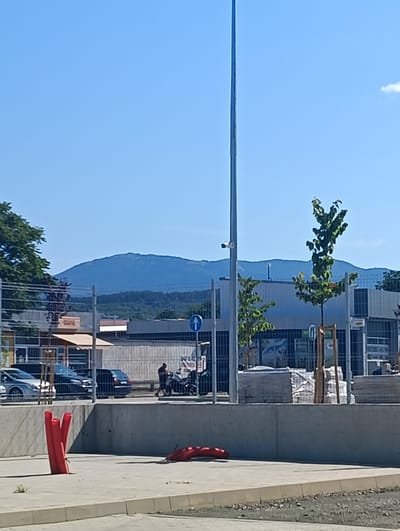 The image captures a scene with a clear blue sky in Kazanlak, Bulgaria. In the foreground, a paved area features several bright red, cylindrical objects protruding from the ground, likely bollards or markers. A long, red, curved object lies on the ground to the right. A grey concrete wall runs horizontally across the middle ground, separating the foreground from the background.
Beyond the wall, a street or parking area is visible, with several cars parked. One silver car is on the left, and a dark-colored hatchback is to its right, followed by another dark car. A person is walking near a motorcycle on the right side of this area. A tall, thin metal pole, possibly a lamppost or flag pole, stands prominently in the center of the image, extending from the foreground into the sky. Adjacent to this pole are two young trees with green leaves.
Further in the background, a low-rise building complex with reflective windows and signage is present. A blue directional sign with a white arrow pointing upwards is affixed to a pole. A chain-link fence borders this building area. In the far background, a large, hazy mountain range dominates the horizon, its slopes covered with dense forest. The overall scene appears to be an outdoor urban or semi-urban environment on a sunny day.