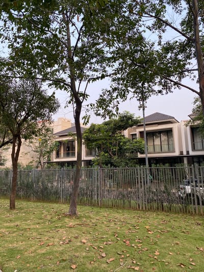 The image depicts a residential street in North Jakarta, Indonesia, likely in a modern housing complex. The foreground features a well-maintained lawn scattered with fallen brown leaves, indicating an autumn or dry season. Two trees stand on the left and right sides of the lawn, their branches extending upwards, with the one on the left being taller and more slender than the one on the right. A grey metal fence separates the lawn from the property behind it.
Beyond the fence, a row of contemporary houses is visible. These houses are characterized by their sleek design, featuring cream-colored facades, large windows with dark frames, and dark-tiled roofs. One house in the center showcases a prominent, multi-story window. Mature trees and lush green foliage are interspersed among the houses, suggesting a well-landscaped environment. A tall, slender street lamp or utility pole stands in front of one of the houses. The sky above is a pale, overcast grey. No people are visible in the scene.