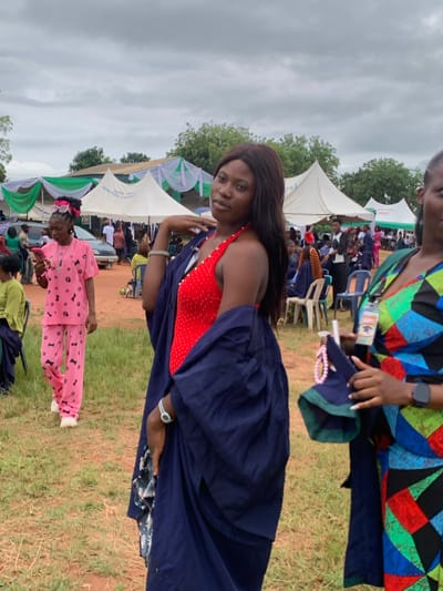 The image shows an outdoor event in Ukwu-Abwa, Nigeria. A woman in a red dress and a dark blue gown stands in the foreground. Another woman with a colorful geometric-patterned top stands partially in the upper right corner, holding a beaded necklace and an identification card. In the background, there are tents decorated with green and white fabric, under which people are seated. A child in pink pajamas with paw print patterns walks in the foreground as well. The ground is covered in dry grass, and trees are visible in the background. The sky is overcast.