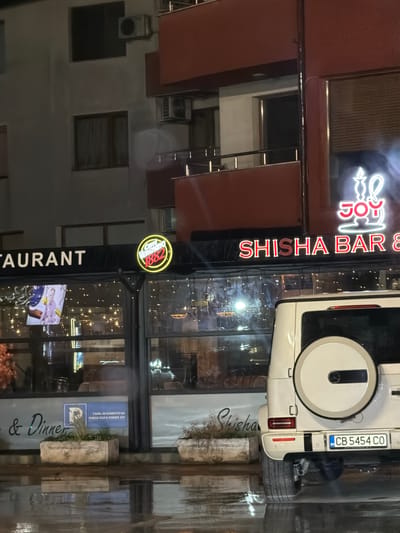 This detailed image captures a nocturnal street scene, positioned at the ground level of a multi-story building. As instructed, the scene is located in the city of Haskovo, Bulgaria.
The building features a light-colored, possibly off-white or light grey, facade on its upper floors, with dark, unlit windows. Several external air conditioning units are mounted on the exterior walls of the upper levels. A central section of the building displays dark reddish-brown cladding or balcony structures.
The ground floor accommodates commercial establishments. On the left, a sign partially visible reads "RESTAURANT". Adjacent to this, a circular, yellow neon sign prominently displays "Caffè Vergnano 1882". To its right, a red neon sign with a stylized hookah emblem illuminates "JOY SHISHA BAR &". Below this signage, a rectangular sign visible through the glass storefront, written in Bulgarian, states "САМО ЗА КЛИЕНТИ НА SHISHA BAR & DINNER JOY" (FOR CLIENTS ONLY OF SHISHA BAR & DINNER JOY) and includes a blue parking symbol. The interiors of these establishments are illuminated by warm-toned string lights and internal fixtures, with seating visible through the windows. Planters containing sparse vegetation are positioned on the sidewalk in front of the storefronts.
A white Mercedes-Benz G-Class SUV is parked with its rear oriented towards the viewer. The vehicle is equipped with a spare tire mounted on its rear door. The Bulgarian license plate, "BG CB 5454 CO", is clearly legible; the "CB" prefix denotes the Sofia City region.
The ground surface is visibly wet and highly reflective, suggesting recent rain or damp conditions. Prominent reflections of the vehicle, neon signs, and interior lights are cast upon the wet pavement. The overall lighting scheme indicates a nighttime or late evening environment. No individuals are present or discernable within the captured frame.