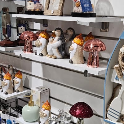 This is an indoor, slightly askew, eye-level photograph depicting shelves displaying various decorative items in a store.
The shelves are white with metal support brackets. The top shelf holds a blue and white boat-shaped ornament filled with rope and a white paddle. Below are several shelves with various items. One shelf features decorative gnomes and mushroom-shaped decorations covered in bronze mosaics. There are also vintage-style microphones and other small items with price tags. Another shelf shows a green soap dispenser, more gnomes, and what appears to be packaging labeled "HOME & STYLING COLLECTION" and "HOURGLASS". Additional items such as boxes and labels are visible on other shelves.
The environment appears to be a retail space with bright, even lighting. The photograph is shot in a manner that suggests the viewer is standing directly in front of the display shelves.
No specific details suggest that the photo was taken in Fez, Morocco.