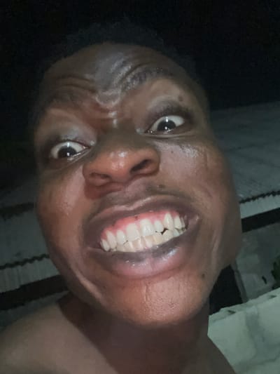 A close-up, low-light photograph captures a person with a dark complexion, looking directly at the camera with an exaggerated facial expression. The subject's forehead is deeply furrowed, indicating strong muscle contraction. Both eyes are wide open, revealing prominent whites and dark pupils, with distinct reflections from a light source. The nose is broad. The mouth is open in a wide, intense smile or grimace, fully exposing a set of uniformly white upper and lower teeth and visible pink gums. The skin appears somewhat moist or reflective under the lighting. The background is dark and out of focus, suggesting an evening or night setting. Faint, dimly lit structural elements are discernible on the right side, including what appears to be corrugated metal sheeting and lighter-colored, uneven surfaces, possibly concrete or plaster walls, consistent with an outdoor or semi-outdoor residential area within the city of Ikorodu, Nigeria.